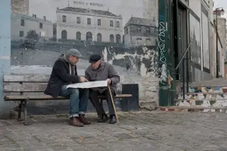 Two people on a bench in Montreuil studying an old map together, with subtle urban historical details around them in soft daylight.
