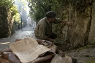 Older craftsman studying medieval stonework on a quiet Montreuil street, morning light, manuscript visible in foreground