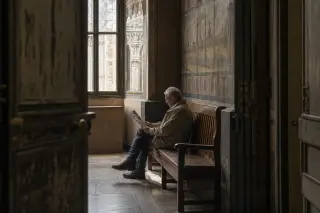 Elderly man reading a heritage brochure alone on a bench in a dimly lit historic hallway in Montreuil, sunlight streaming through tall windows, viewed discreetly from behind a half-open door.
