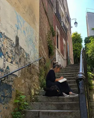 Middle-aged woman sketching quietly on a sunlit, weathered outdoor staircase between old brick buildings in Montreuil, surrounded by faded murals and greenery.