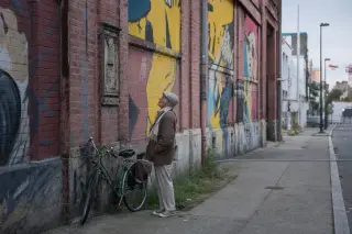 Older resident observing a faded industrial brick building with street art in Montreuil, soft daylight enhancing urban textures.