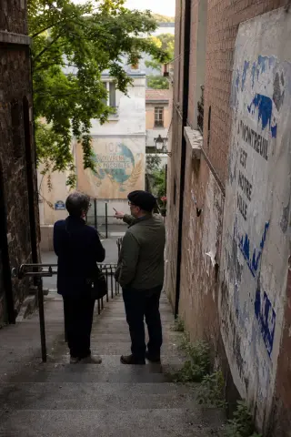 Two people conversing naturally on a worn urban staircase in Montreuil, beside brick walls and a faded mural in soft afternoon light.