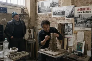 Two people in a Montreuil artist studio, one sculpting clay, the other observing, surrounded by historic photos and modern posters in soft natural light