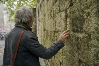 An elderly craftsman studies a mossy medieval wall in Montreuil, softly lit by natural daylight, suggesting quiet reflection on the town's changing history.