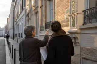 Two people observe architectural details on a Montreuil building façade in soft morning light, absorbed in conversation, with urban textures and greenery around them.