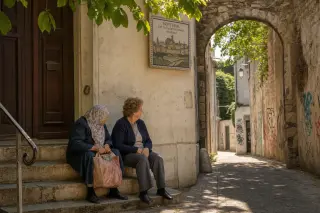 Two elderly women quietly seated on worn stone steps under a tree in Montreuil, with graffitied walls and hints of historic architecture in soft midday light
