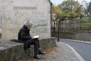 Elderly woman sketching on a stone wall near mossy historic facade and the Murs à pêches in Montreuil, under gentle daylight.